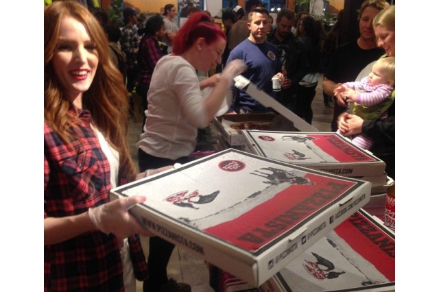 Two woman handing out pizza slices from a stack of red and white pizza boxes to a crowd indoors at an office party.